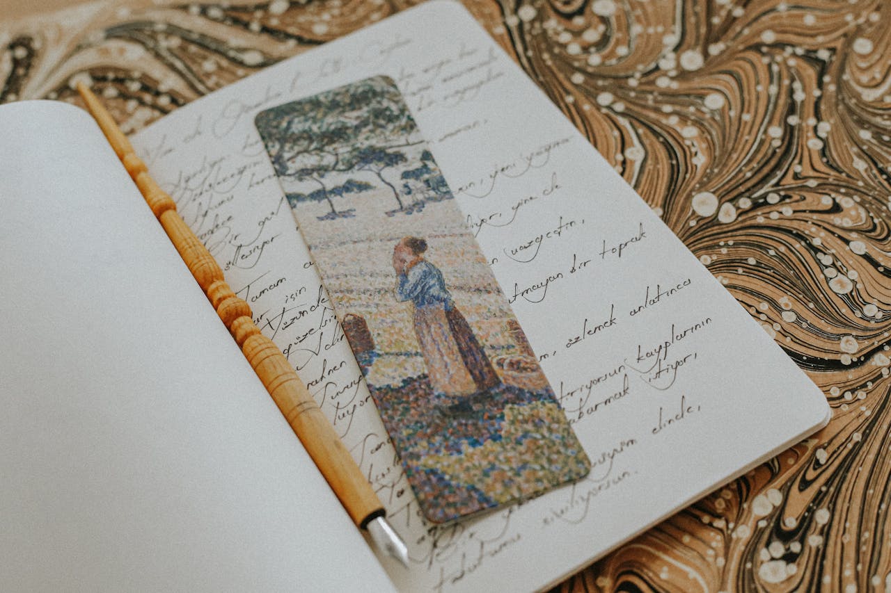Open notebook with handwriting, a painted bookmark, and a pen on a marbled surface.
